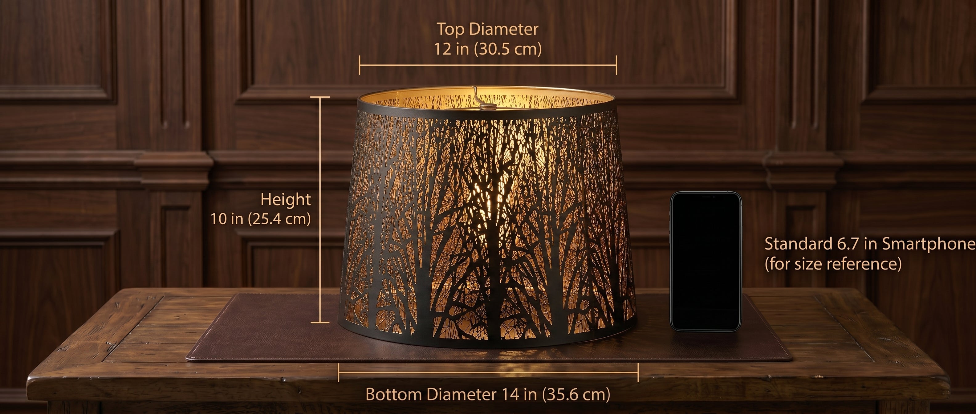 Large Forest Etched Metal Lamp Shade Precise Dimensions Size Reference Chart - Shade Resting on Brown Leather or Faux Leather Placemat on Rustic Dark Wood Plank Table or Desk with Wood Grain and Joinery Details, Rich Dark Walnut Wood Panel Wall Background with Decorative Panel Moldings - Precise Measurements Marked with Gold Lines: TOP DIAMETER 12 in (30.5 cm), BOTTOM DIAMETER 14 in (35.6 cm), HEIGHT 10 in (25.4 cm) - Size Comparison with Standard 6.7 in Black Smartphone (Modern iPhone Style) Placed to Righ