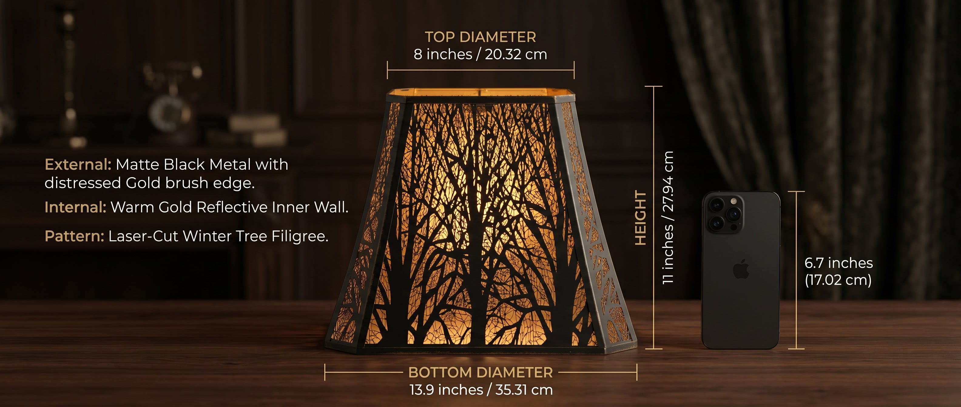 Large Rectangle Forest Etched Metal Lamp Shade Size and Material Specifications - Product Features: EXTERNAL - Matte Black Metal with Distressed Gold Brush Edge, INTERNAL - Warm Gold Reflective Inner Wall, PATTERN - Laser-Cut Winter Tree Filigree, Dimensions - TOP DIAMETER 8 inches (20.32cm), BOTTOM DIAMETER 13.9 inches (35.31cm), HEIGHT 11 inches (27.94cm) - Size Comparison with Standard 6.7 inches (17.02cm) Black iPhone Smartphone - Shade shown illuminated on Polished Dark Wood Table with Vintage Brass Ca