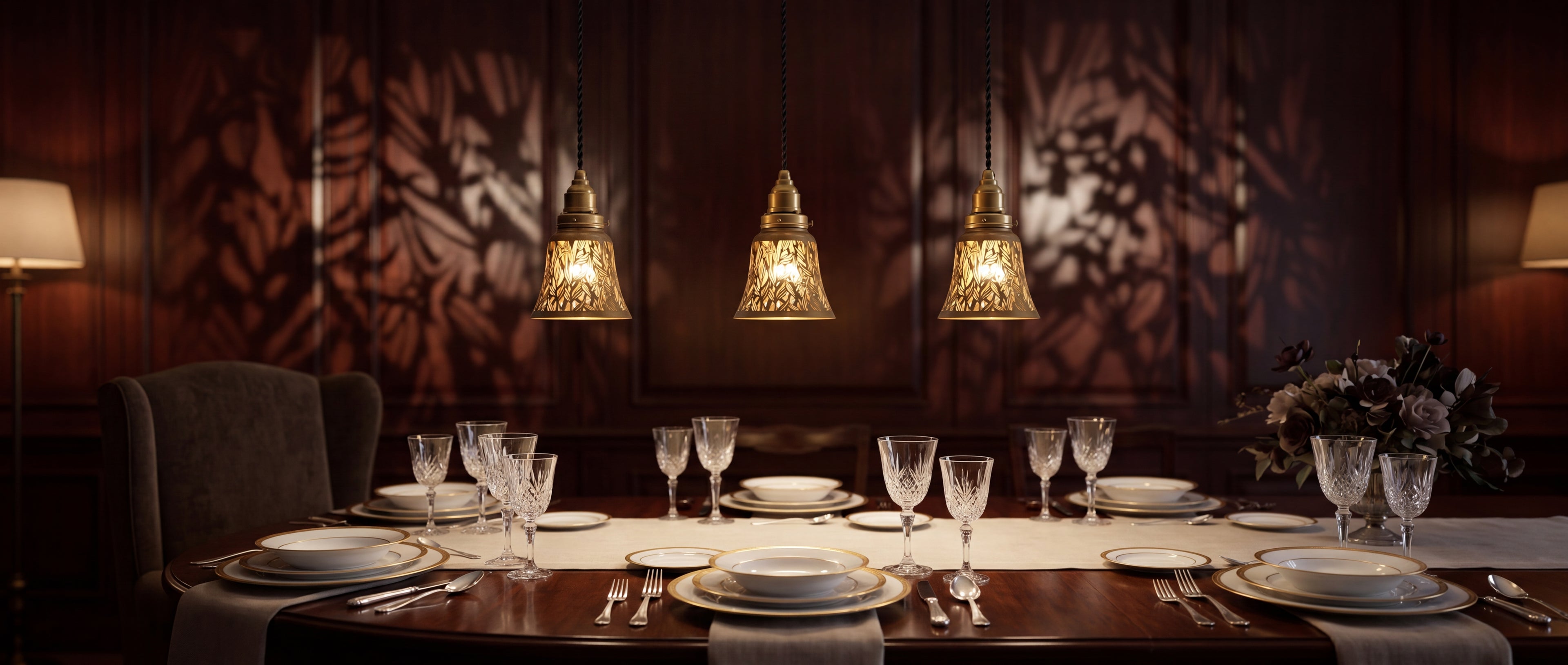 Laser-cut starburst metal lamp shade set of 4 pack clip-on small decorative shades, three pendant hanging lights in row over dining room table, antique brass finish gold interior botanical leaf pattern creating warm ambient light projection with intricate shadow pattern on dark wood panel wall, elegant dining table set with crystal stemware wine glasses white dinner plates and silverware on ivory linen table runner with dried flower floral arrangement, ideal lamp shade replacement for pendant lights chandel