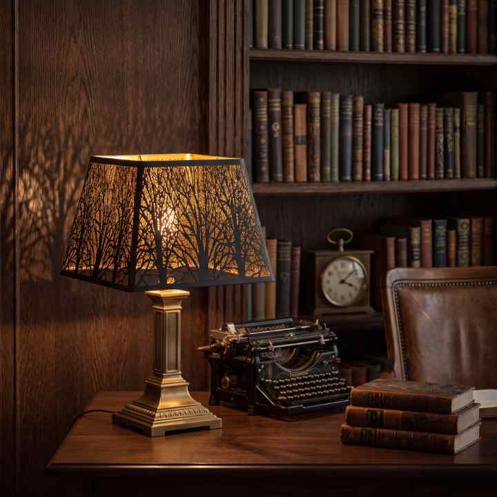 Modern softened square metal lamp shade on vintage brass table lamp, black matte exterior gold interior with subtle woodland tree texture, warm ambient lighting in industrial minimalist study office