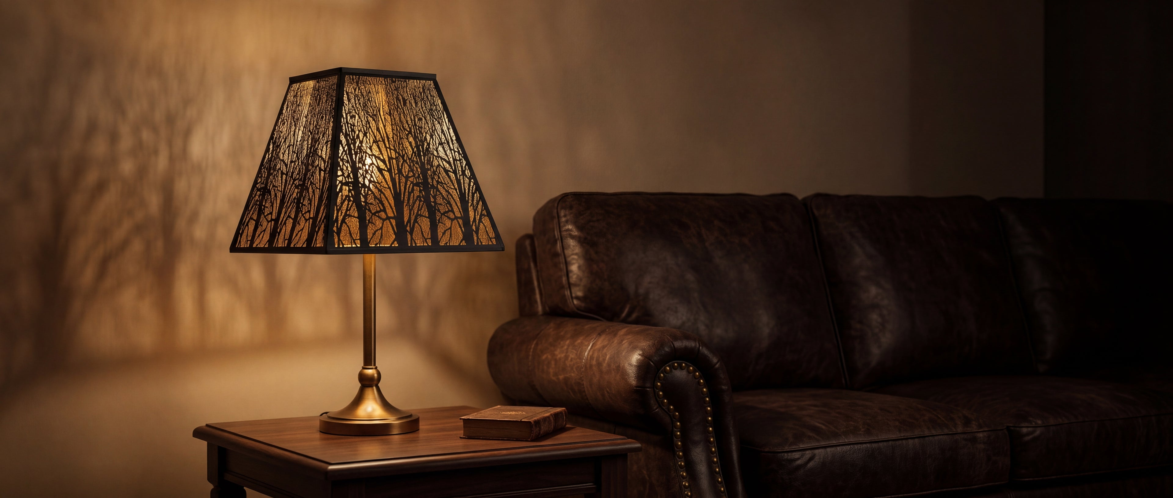 Pyramid Metal Lamp Shade with Square Bell Shape and Forest Silhouette Design Installed on Elegant Antique Brass or Antique Gold Candlestick Table Lamp with Decorative Urn-Shaped Stem and Round Fluted Base - Lamp Resting on Dark Wood Side Table or End Table with Decorative Stack of One Antique Leather-Bound Book with Brown or Tan Cover - Elegant Traditional or Farmhouse Living Room Setting Features Rich Brown or Cognac Chesterfield Tufted Leather Sofa or Couch with Brass Nailhead Trim - Beautiful Intricate F